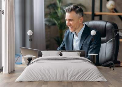 Influential successful Caucasian adult businessman or manager in a formal suit uses a laptop, communicates via video conferencing, online meetings, sits office, types on a keyboard, smiles friendly Wall mural