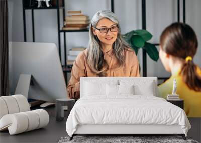 Intelligent confident influential gray-haired female asian wearing glasses, business lady, HR manager, talking to job candidate in modern office, holds and studies resume, smiling friendly Wall mural