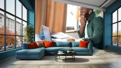 Joyful African American guy dressed in stylish clothes uses a laptop for online work, sitting at his desk, happily looks at the screen, got a good message Wall mural