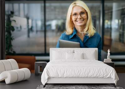 Joyful blonde-hair business woman in eyeglasses is looking at the camera, holding laptop in arms and smiling. Beautiful middle aged lady in stylish wear stands in modern office Wall mural