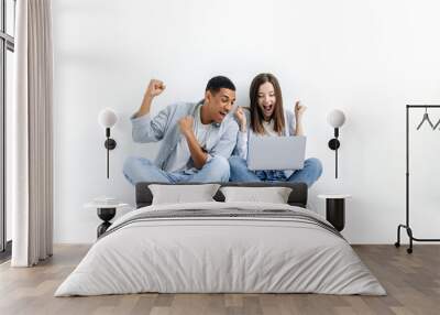 Joyful excited man and woman of different nationalities, sit on the floor on a white isolated background with a laptop, look amazed at the screen, rejoice at the good news, win, success, smile happily Wall mural