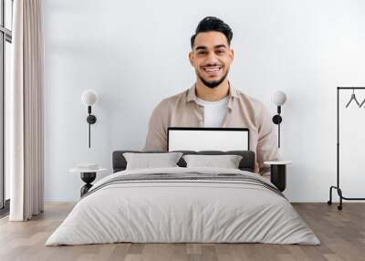 Joyful handsome Indian or Arabian young man, student or freelancer, stands on white isolated background, holding open laptop with blank white mockup screen for presentation, looking at camera, smiling Wall mural