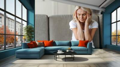 Middle aged blonde woman sits on couch at living room holding her head with her hands, feels unhappy because of headache, personal troubles, illness or bad news, she need psychological or medical Wall mural