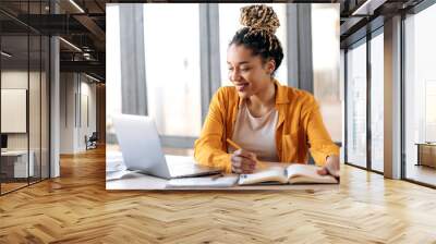 Online education, e-learning. Happy african american young woman in stylish casual clothes, studying remotely, using a laptop, listening to online lecture, taking notes while sitting at home, smiles Wall mural