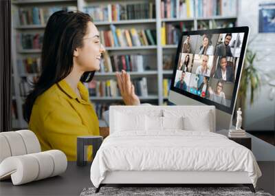 Online education. A female student learning online at home, she waving hand to other students or colleagues on computer monitor in video conference and smiling Wall mural