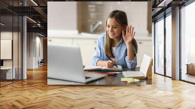 Online lesson, distance learning, homeschooling during quarantine. Caucasian pretty schoolgirl at online lesson by video call waves hand, greeting teacher, at home using laptop, sitting at the work Wall mural