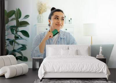 Online shopping concept. Joyful caucasian woman using laptop computer with credit card making online payment, sits at a desk, thoughtfully looks away, thinking about what to spend the money on, smiles Wall mural