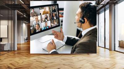 Online video conference. Successful young adult businessman or manager with headphones discuss with colleagues by video call about financial graphs and strategy and another working moments Wall mural