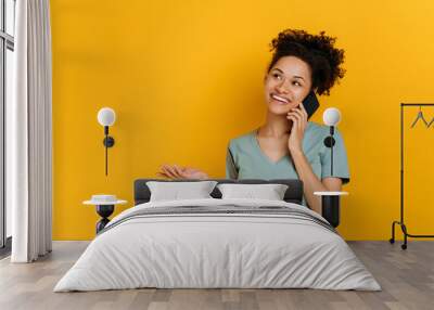 Phone talk by mobile phone. Happy excited african american young woman has pleasant phone conversation, happy facial expression, stands on isolated orange background, looks away, gesturing hand, smile Wall mural