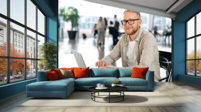 Photo of a positive attractive clever caucasian business man, with glasses, executive, programmer, company seo, manager, sits at a desk with a laptop in coworking center, looks at camera, smiling Wall mural