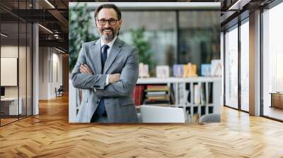 Portrait of a confident middle aged entrepreneur in stylish suit and eyeglasses standing in modern office, looking at the camera and friendly smiling Wall mural