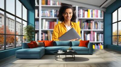 Portrait of a girl student. An african american pretty, smart confident female student, stylishly dressed, holding a laptop, standing in the university library, looking at the camera, smiling friendly Wall mural