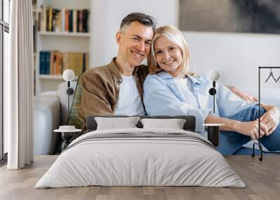 Portrait of a pleasant happy mature caucasian married couple relaxing on sofa in living room at home, spending time together, dressed in casual stylish clothes, smiling at camera, happy together Wall mural