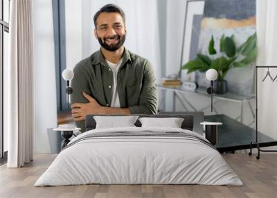 Portrait of a successful confident Indian man, creative manager, IT specialist, designer, wearing casual wear, standing near desk in the office with arms crossed, looks at the camera, friendly smiling Wall mural