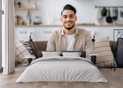 Portrait of an attractive handsome indian or arabian guy in a stylish casual wear, sitting on sofa at home in a living room, in a stylish interior, looking at the camera, smiling positively Wall mural