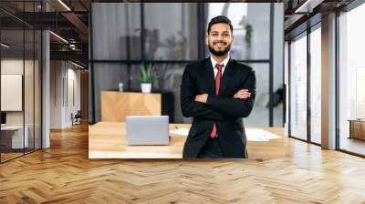 Positive handsome confident successful Indian or Arabian businessman, in a suit, male entrepreneur, stands in a modern office near the desktop with crossed arms, looks at the camera, smiles friendly Wall mural