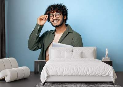 Smart handsome positive indian or arabian millennial guy, with glasses, student or freelancer, holding a laptop in hand, standing on isolated blue background, looking at the camera, smiling friendly Wall mural