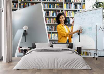 Stylish African American female coach, stands at whiteboard, in casual clothes, uses computer, leads an online lesson, communicates with students via video conference, looks at screen,distant learning Wall mural