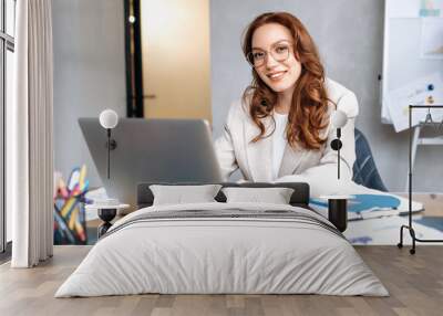 Successful redhead business woman is using laptop, working on new project in modern office. Stylish beautiful female employee in eyeglasses is looking at the camera, smiling Wall mural