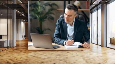 Successful serious adult attractive caucasian business leader or ceo, sitting at workplace in the office in a formal suit, concentrated working in a laptop, browsing internet, analyzes information Wall mural