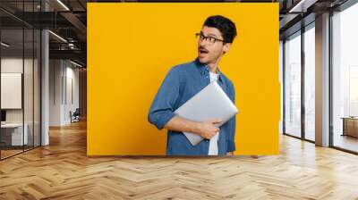 Surprised shocked caucasian stylish smart guy with glasses, is holding laptop in hand, is looking back with amazement looking towards empty space, standing on isolated orange background Wall mural