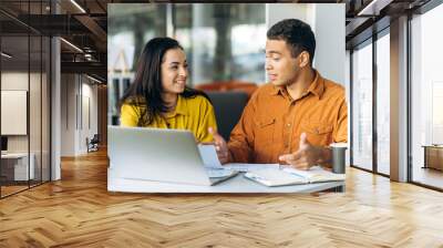 Two joyful multiracial colleagues or students, caucasian girl and hispanic guy, are using a laptop while sitting at workplace discussing about job or education and smiling Wall mural