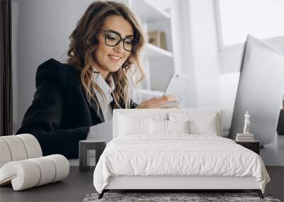 Young attractive business woman working in her office with a laptop, looks into the phone and smiling Wall mural