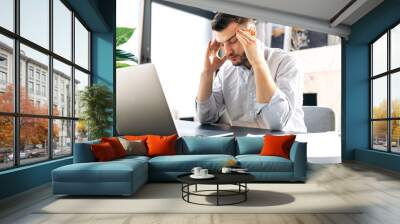 Young tired caucasian man in a formal shirt sitting at his workplace, holding his head with his hands, closing his eyes from headache due to long work in a laptop Wall mural