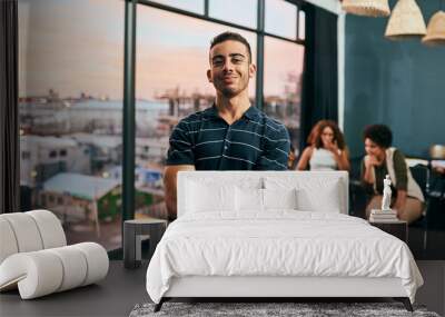 He has big dreams for their small business. Portrait of a cheerful young man standing with his arms folded with his work colleagues grouped together in the background. Wall mural