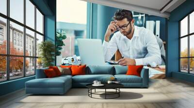 His job is seriously starting to stress him out. Shot of a young businessman looking stressed out while working on a laptop in an office. Wall mural