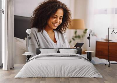 Cheerful black woman resting in bed while scrolling on her phone. Wall mural