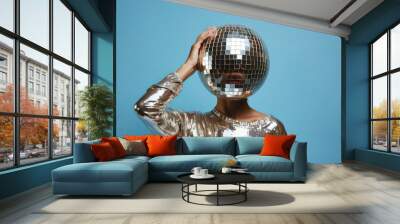 A fashionable woman wearing a silver outfit poses putting a vintage disco ball on her head, set against a blue studio backdrop. Wall mural