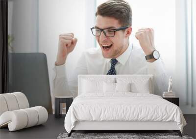 Happy young businessman raising hands in front of laptop at office desk Wall mural