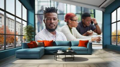 young African American man posing for the camera while sitting at a table in front of his colleagues Wall mural