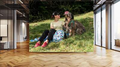 Happy family mother with daughter and two dogs sitting on green grass lawn having fun picnic spending time together hiking in the mountains. People and pets outdoors lifestyle photo candid emotions. Wall mural