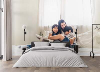 Asian family mother, son and daughter sit on white bed with happiness and smile in bedroom. Young boy and girl hug single mom smiling enjoy playing together at home. Love and warm touch from parent. Wall mural