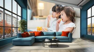 Asian two sister enjoy eating snack on sofa while watching TV at home. Wall mural