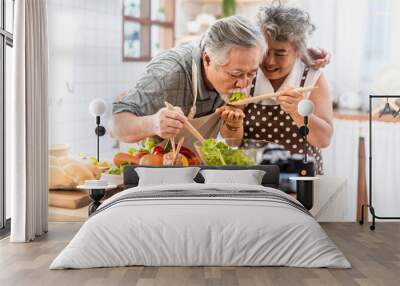 Couple senior Asian elder happy living in home kitchen. Grandfather eating salad dish after cook by grandmother recording vlog vdo for social blogger. Modern lifestyle older people relationship. Wall mural