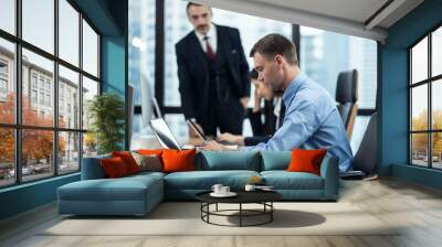 Group of diversity people working in office, two caucasian man and asian woman sitting on desk typing on laptop feeling stress, under pressure by their boss standing behind forcing them complete job. Wall mural