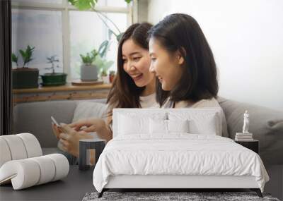 Happy two Asian girls sitting on sofa at home are talking and laughing with smile together while they are looking at the smartphone and using the phone to surfing the internet. Technology concept. Wall mural