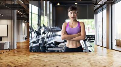 Portrait of young pretty Asian girl with six packs in purple color sportswear standing and crossing arms in gym or fitness club. Behind her is exercise machines and equipment. Fitness & Gym concept. Wall mural