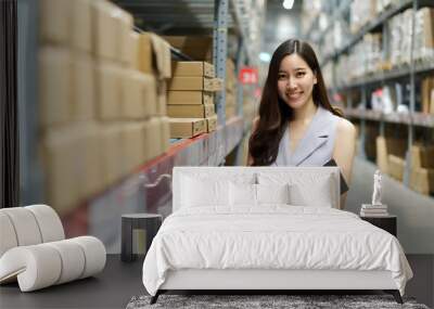 Smart smiling Asian woman working in store warehouse. She is standing and holding document folder and writing some note. She is feeling happy to work. Smart working girl concept. Wall mural