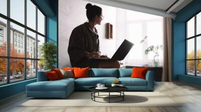 A young woman in home clothes sits with a laptop and looks out the window. Home insulation. Wall mural