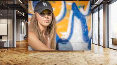 Portrait of young beautiful woman wearing white tank shirt and blue jeans and black hat on brick wall with graffiti background Wall mural