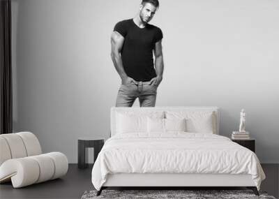 Muscular model sports young man in jeans and black t-shirt on grey background. Black and white fashion portrait of brutal sporty healthy strong muscle guy with a modern trendy hairstyle. Wall mural