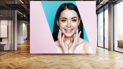 Portrait of model with natural nude make up look into the hole of pink paper. Beautiful young woman with perfect glow clean skin applying moisturizer cream on her face on blue background. Wall mural