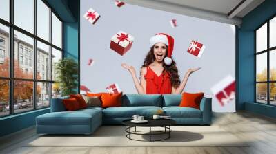 Rain of gifts. Seasonal christmas holidays discounts and sale. Smiling beautiful slim brunette young girl with curly hair in red dress and santa hat expressing happy emotions. Wall mural