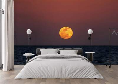 A full moon rising over a dark ocean at sunset, with an orange and red sky in the background Wall mural