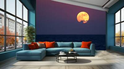 A full moon rising over a dark ocean at sunset, with an orange and red sky in the background Wall mural