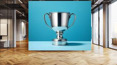 Shiny silver trophy with an elegant base, standing against a blue background. The cup is adorned with intricate designs and features two hands holding the handle at its top. It has been decorated. Wall mural